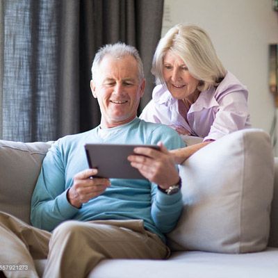 Un couple âgé est assis sur un canapé, les yeux rivés sur une tablette. L'homme la tient, la femme se penche vers lui. Scène de salon.