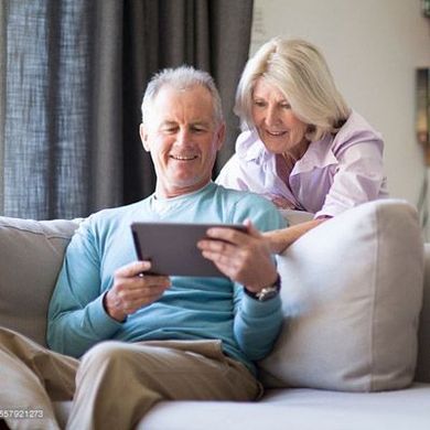 Un couple âgé est assis sur un canapé, les yeux rivés sur une tablette. L'homme la tient, la femme se penche vers lui. Scène de salon.