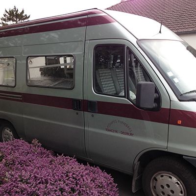 Un camping-car argenté à rayures bordeaux, garé près de fleurs violettes.
