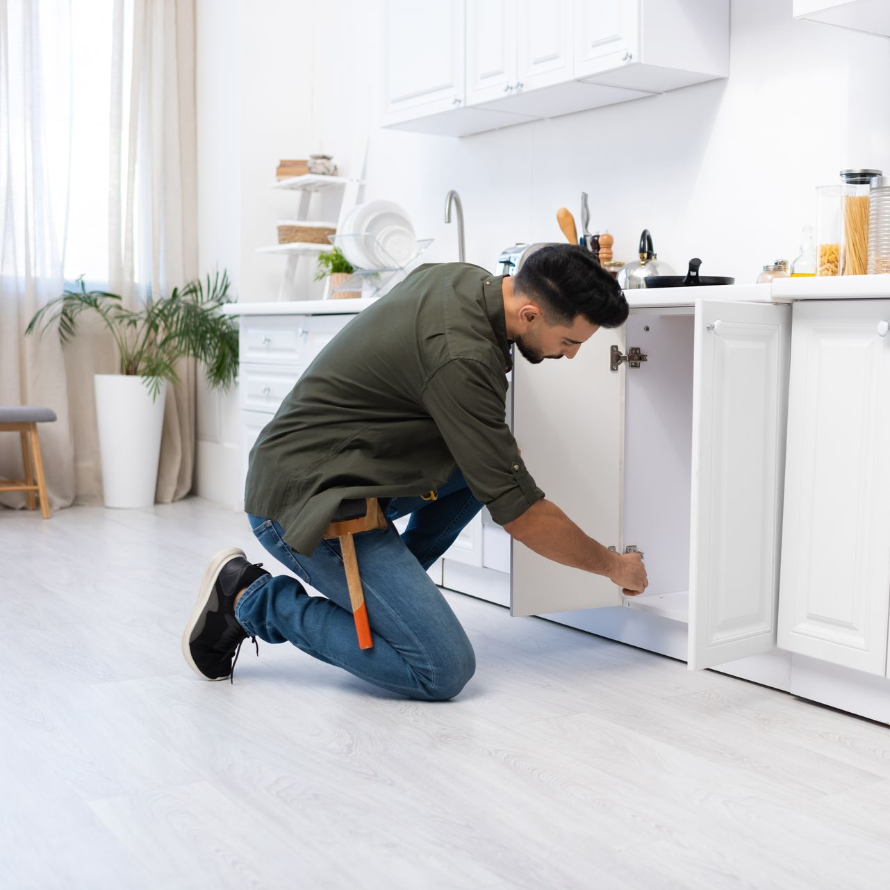 Un homme agenouillé dans une cuisine répare un meuble, portant un jean et une chemise verte.