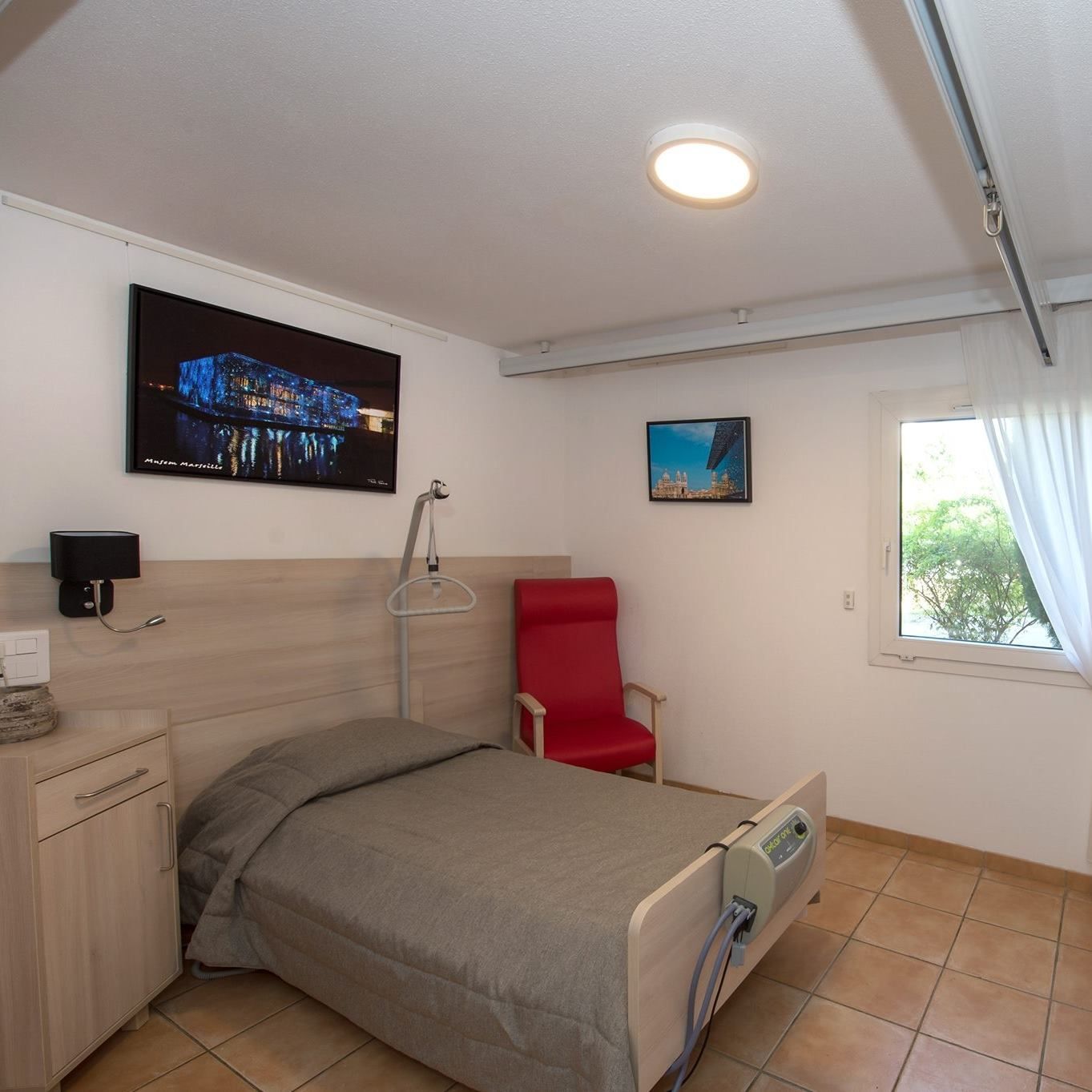 Chambre d'EHPAD moderne avec un lit, une chaise rouge, une télévision murale et une fenêtre avec rideaux.
