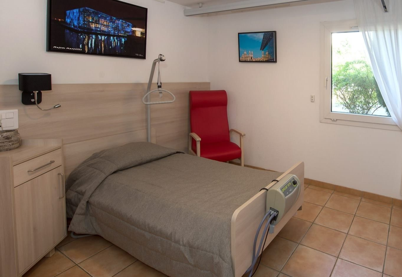 Un lit médicalisé, un fauteuil rouge et une table de chevet dans une chambre d'EHPAD.