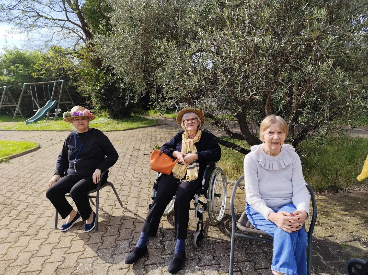Trois personnes sont assises à l'extérieur, sur une zone pavée, devant un grand arbre et des jeux pour enfants.
