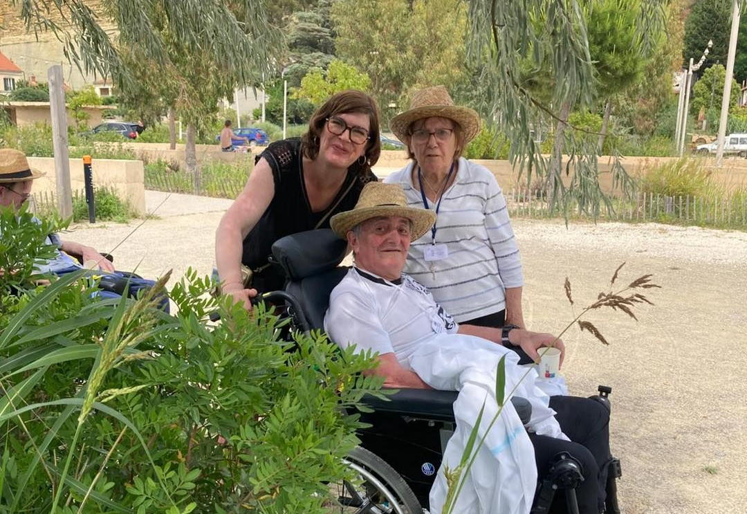 Trois personnes posent en extérieur, l'une d'elles assise dans un fauteuil roulant, avec des arbres en arrière-plan.