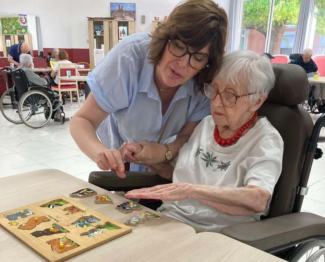 Une aide-soignante assiste une personne âgée en fauteuil roulant qui fait un puzzle en bois.