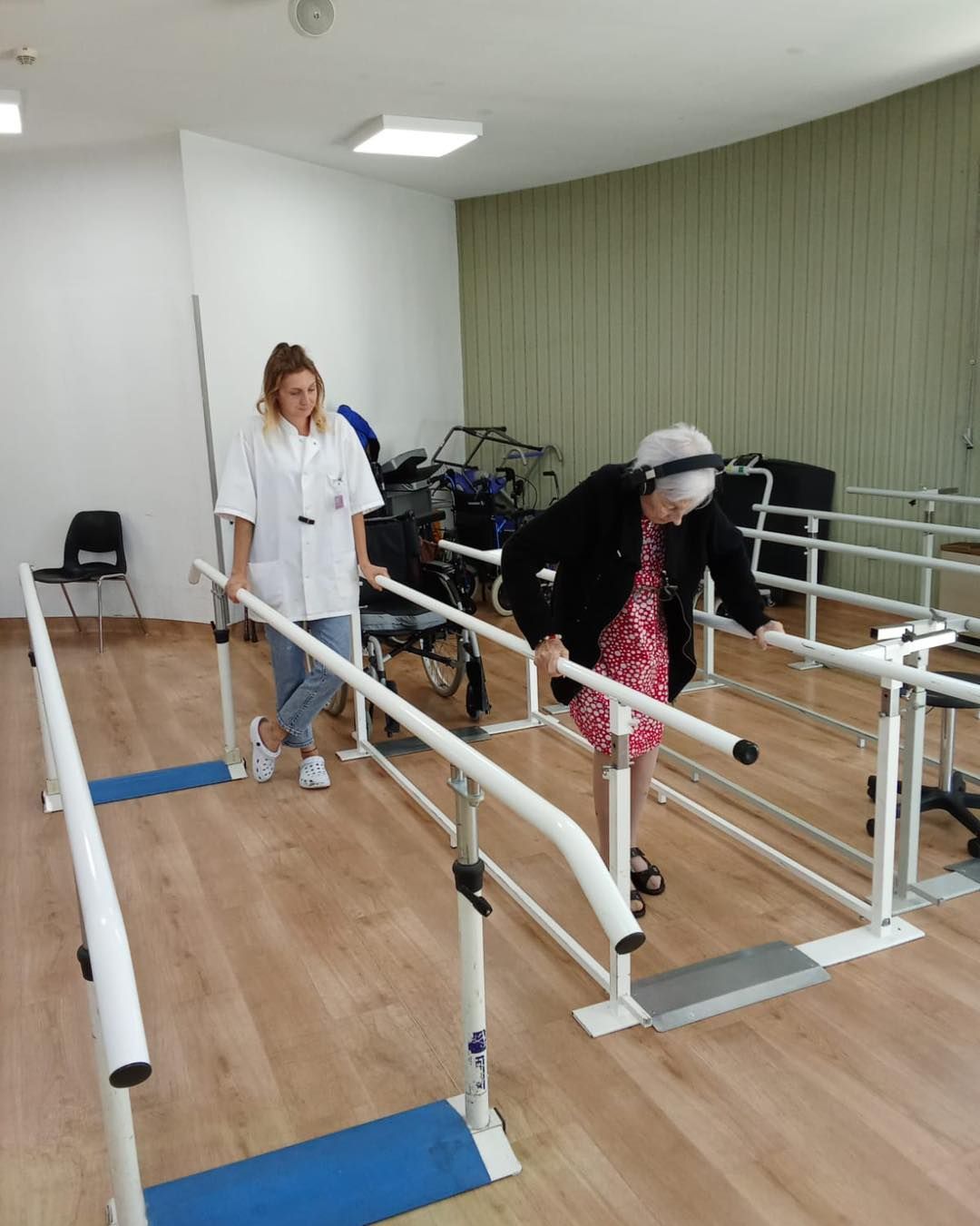 Un physiothérapeute observe une personne s'exerçant à marcher entre des barres parallèles dans une salle de thérapie.