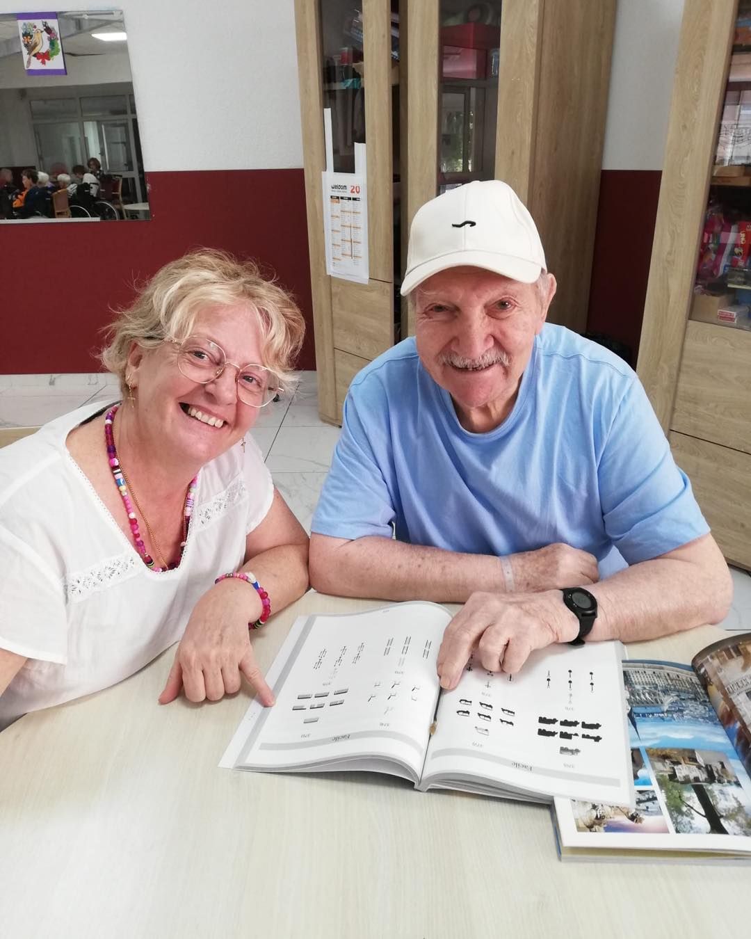 Deux personnes souriantes à une table, regardant un livre ouvert dans un cadre intérieur chaleureux.