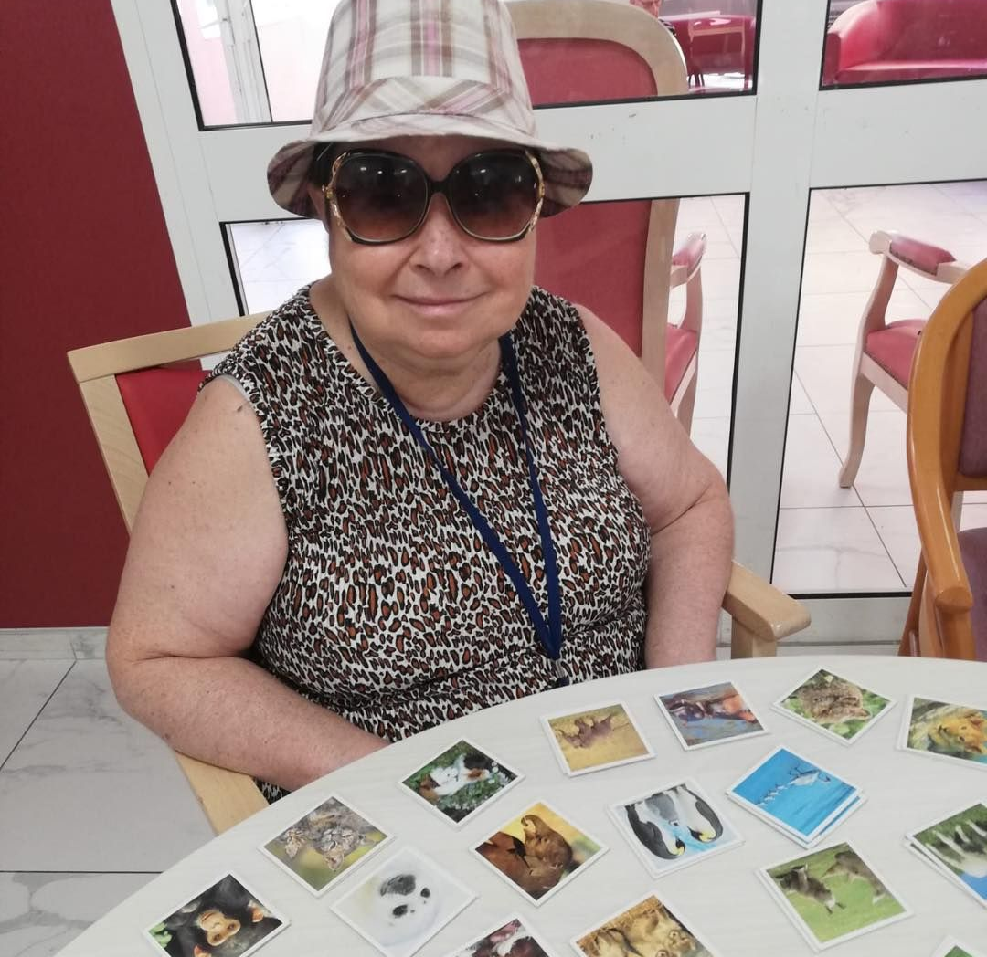 Une femme portant des lunettes de soleil et un chapeau est assise à une table avec des cartes photo dans une pièce lumineuse.