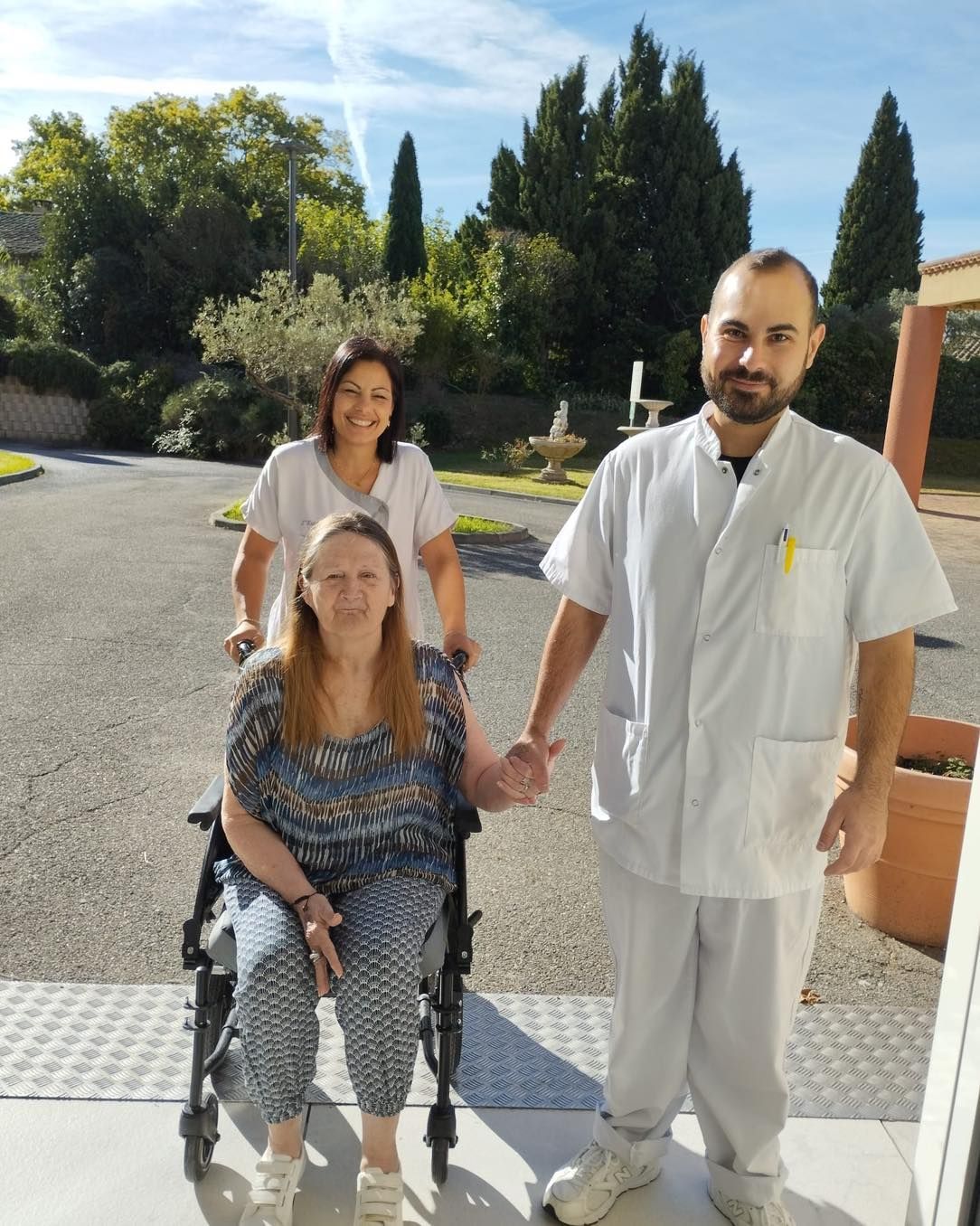 Deux personnes en uniforme blanc se tiennent à côté d'une femme assise à l'extérieur, sur une allée ensoleillée.