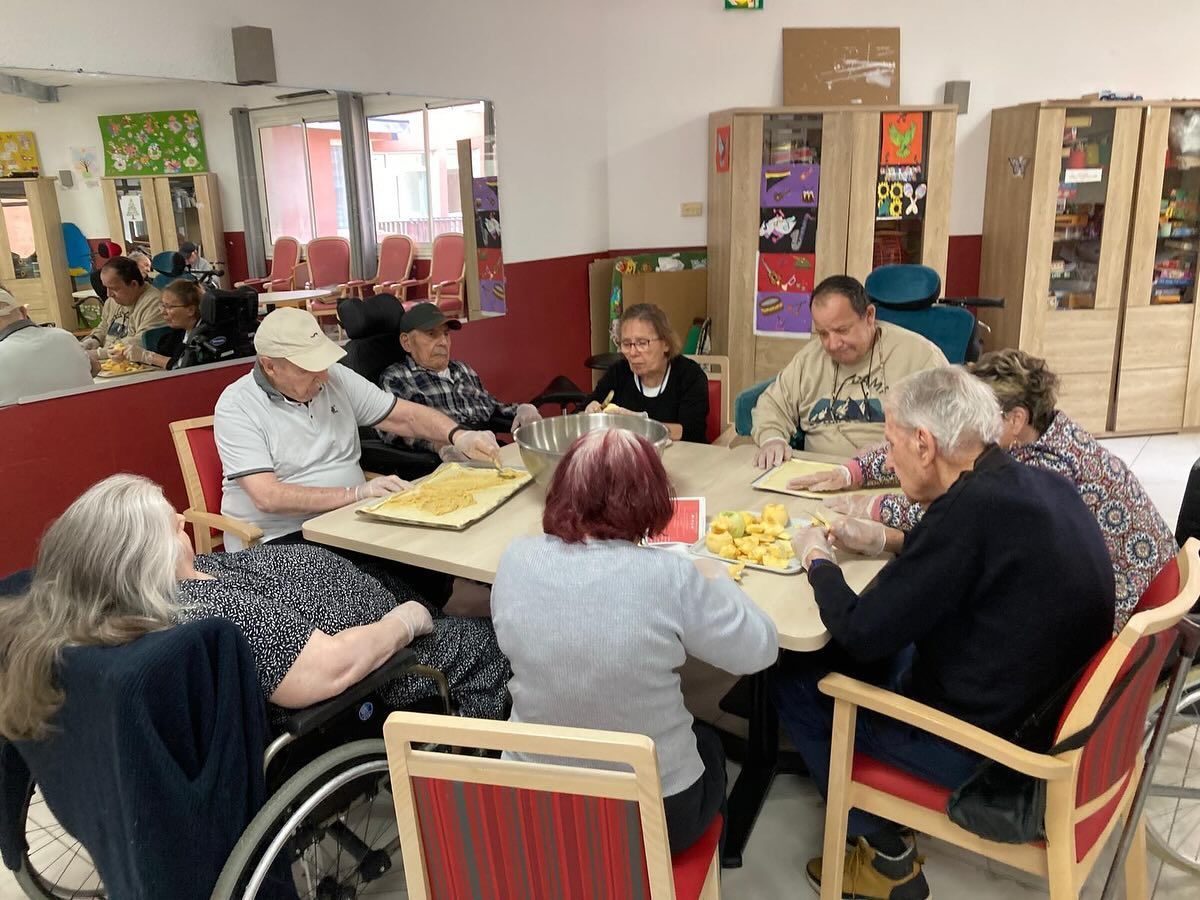Des personnes assises autour de tables dans une salle commune lumineuse discutent et préparent des pâtisseries.