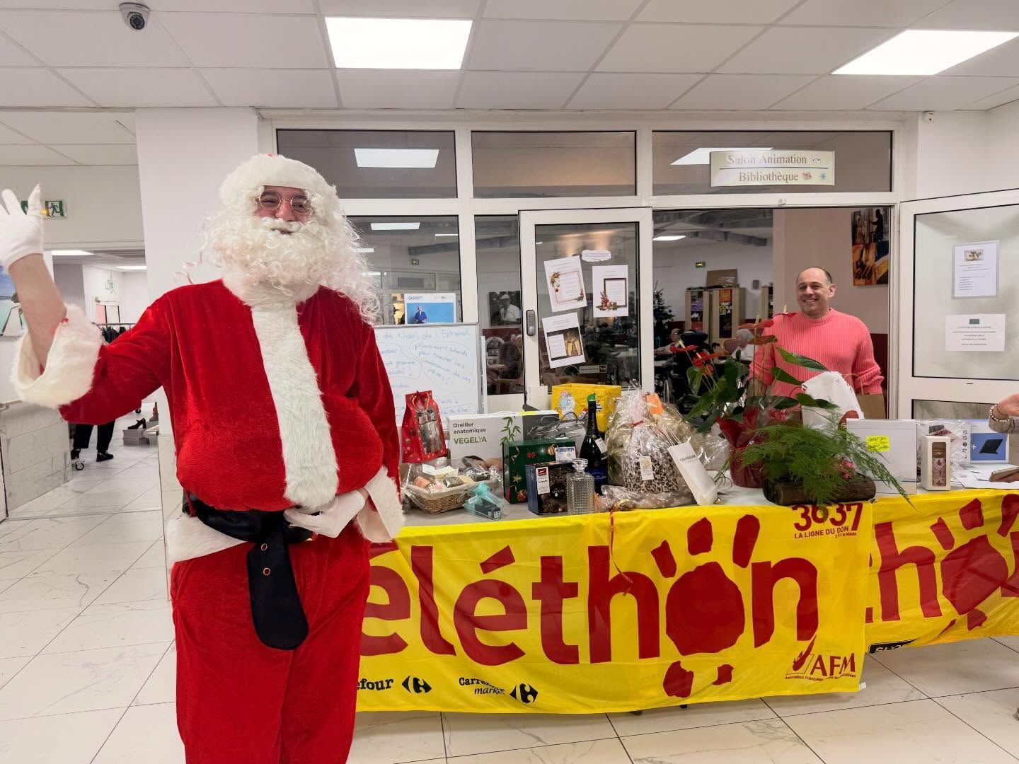Le père Noël fait signe de la main devant une banderole jaune du Téléthon.