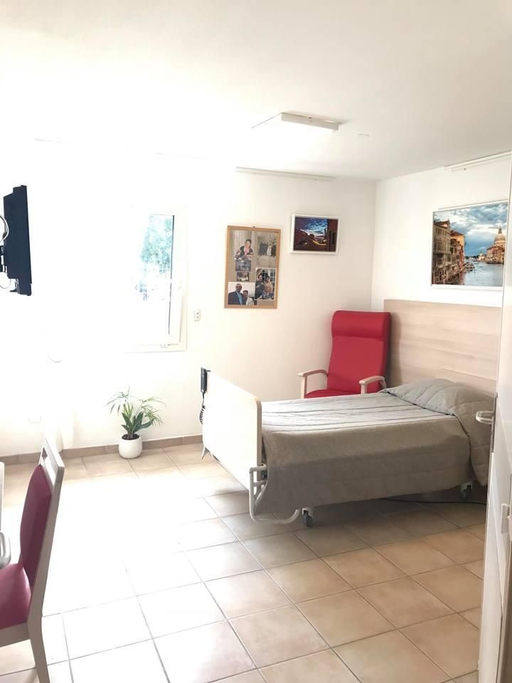 Une chambre d'EHPAD propre et bien rangée, avec un lit simple, un fauteuil inclinable rouge et des décorations murales.