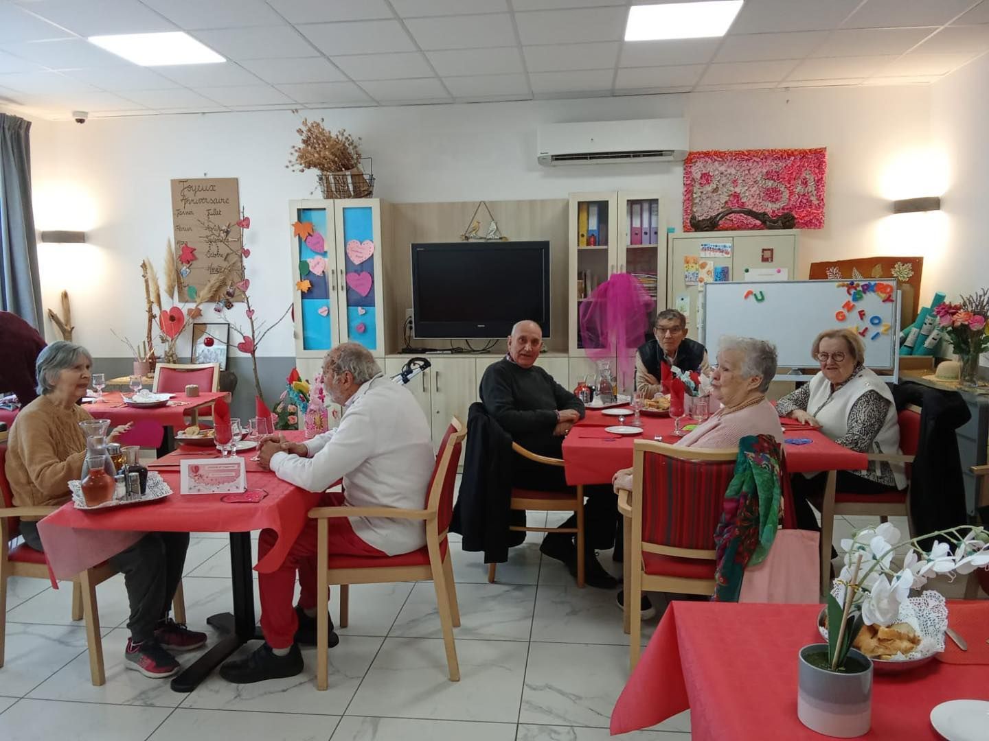Un groupe de personnes âgées est assis autour de tables nappées de rouge, et partage un repas.