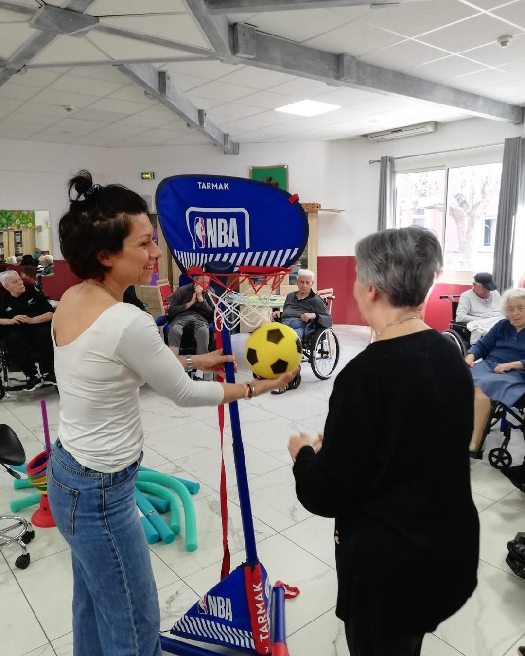 Une animatrice tient un ballon jaune près d'un panier de basket-ball tout en interagissant avec un résident de l'EHPAD.