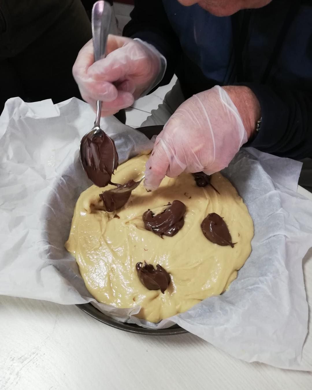 Des mains gantées étalent des noisettes de chocolat sur une pâte à gâteau ronde recouverte de papier sulfurisé.