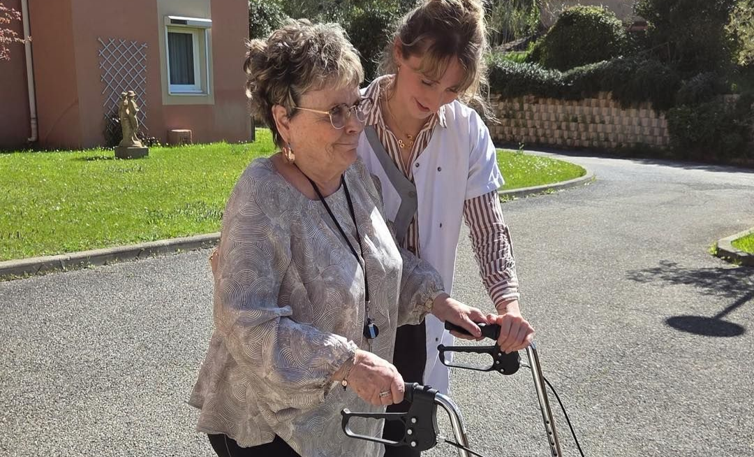 Une aide-soignante accompagne une femme âgée qui marche à l'extérieur avec un déambulateur.