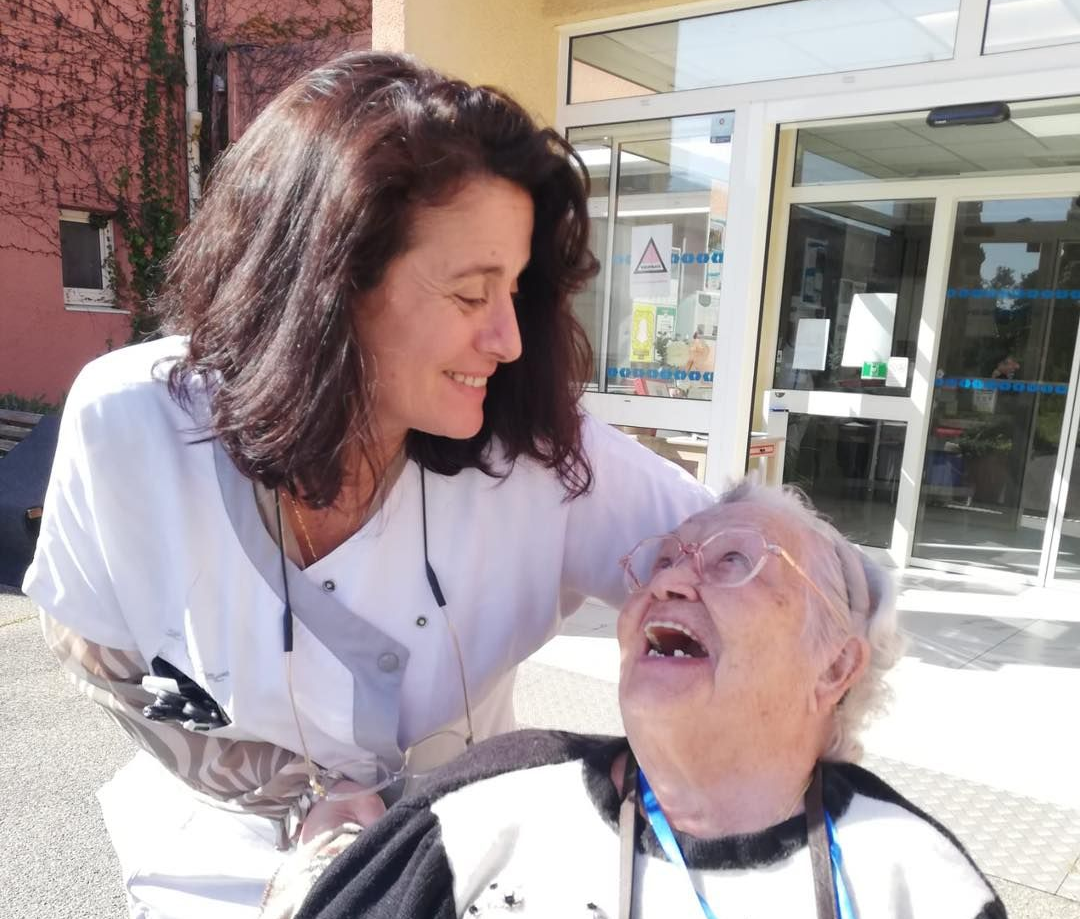 Une infirmière sourit à un patient âgé en fauteuil roulant devant un bâtiment.