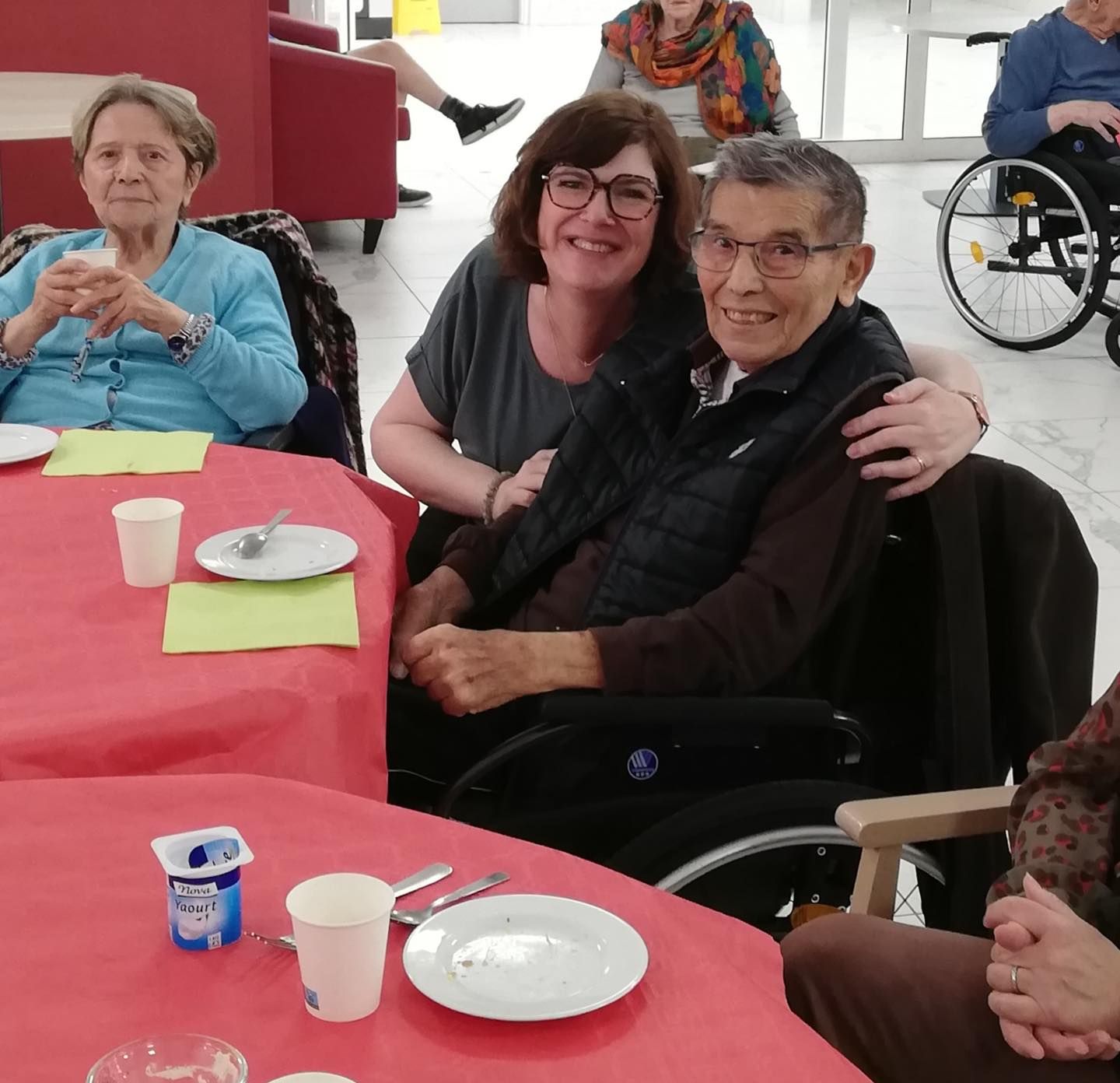 Trois personnes assises à une table recouverte d'une nappe rouge, souriantes dans une salle à manger communautaire.