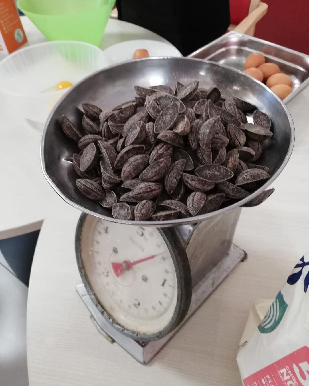 Des morceaux de chocolat empilés sur une balance en métal, avec des œufs et des ustensiles de cuisine en arrière-plan.
