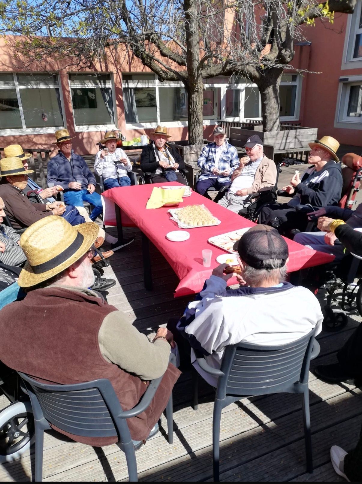 Des personnes assises à l'extérieur autour d'une nappe rouge, échangeant des propos dans une cour ensoleillée.