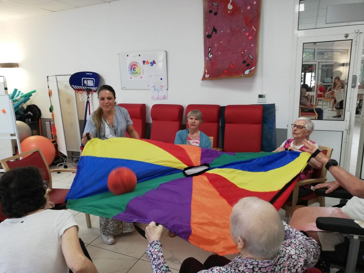 Des personnes assises autour d'un parachute arc-en-ciel dans une pièce lumineuse, participant à une activité de groupe