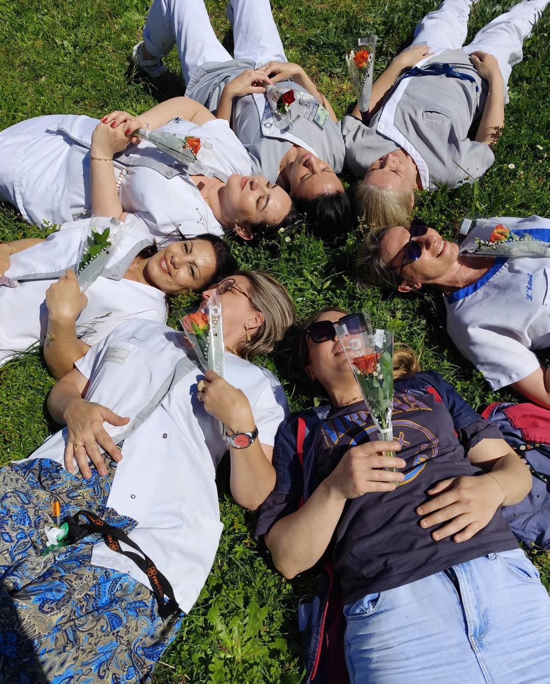 Des aides-soignantes sont allongées dans l'herbe, vêtues de chemises blanches, souriantes et tenant des fleurs.