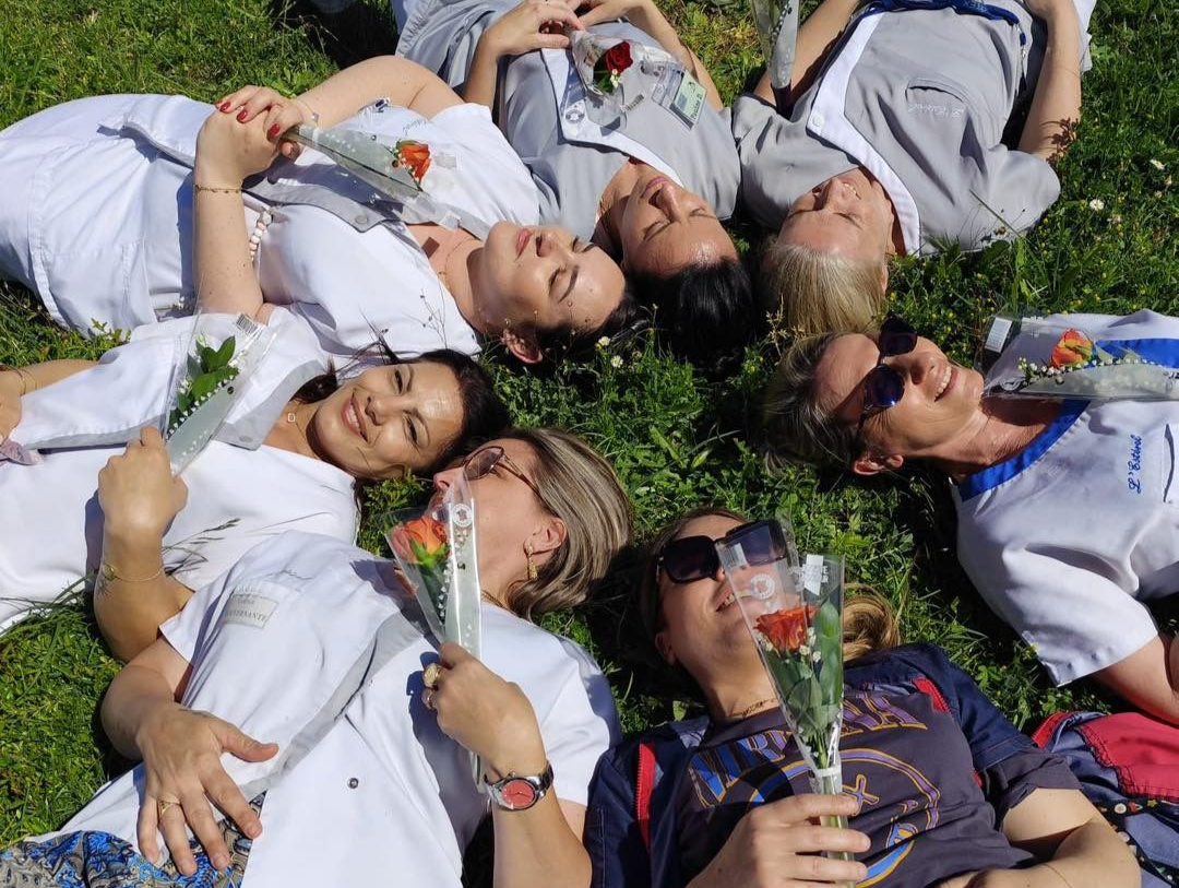 Des aides-soignantes sont allongées dans l'herbe, vêtues de chemises blanches, souriantes et tenant des fleurs.