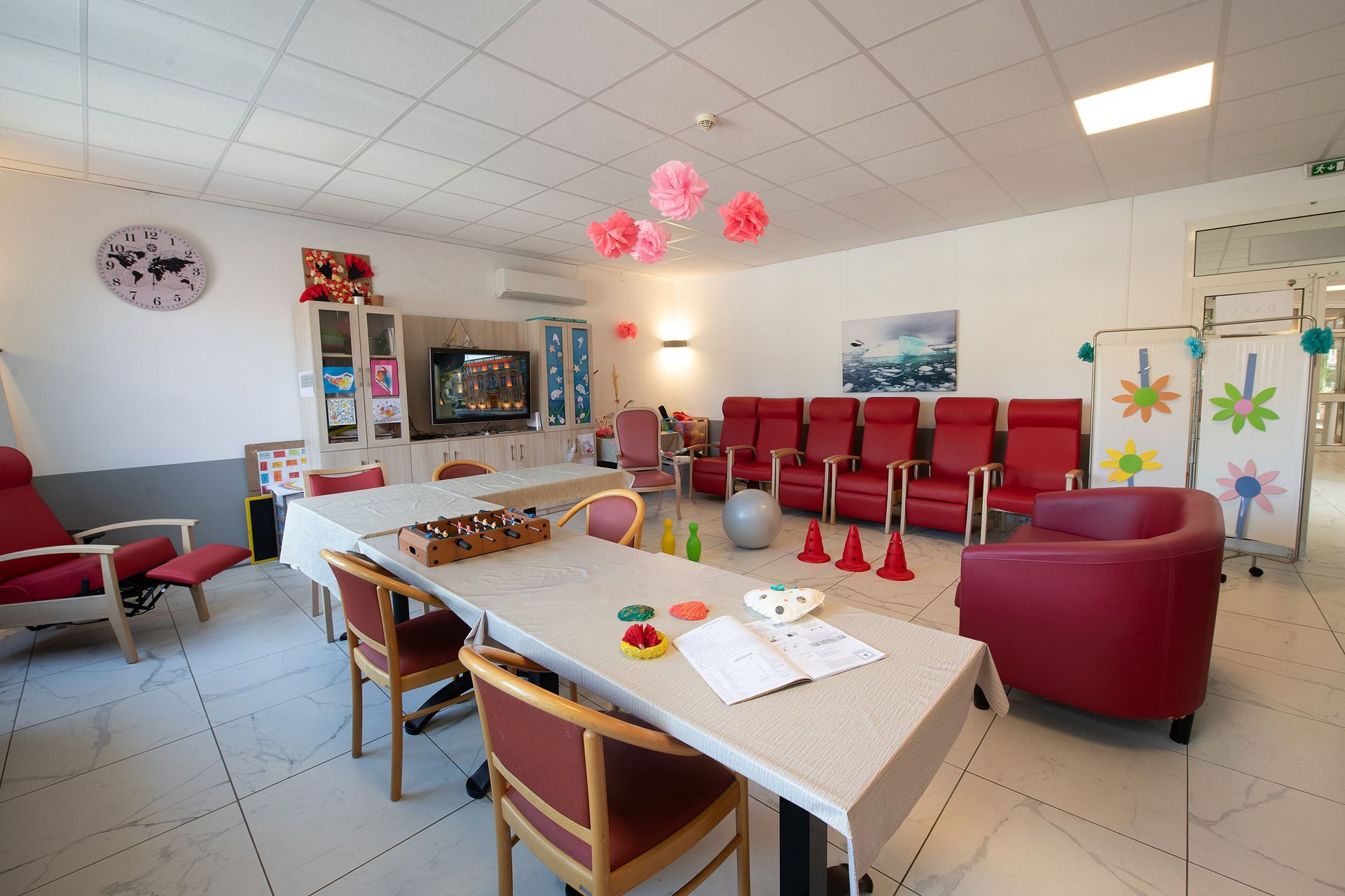 Salle de jeux lumineuse avec chaises rouges, tables, jouets et décorations murales colorées.