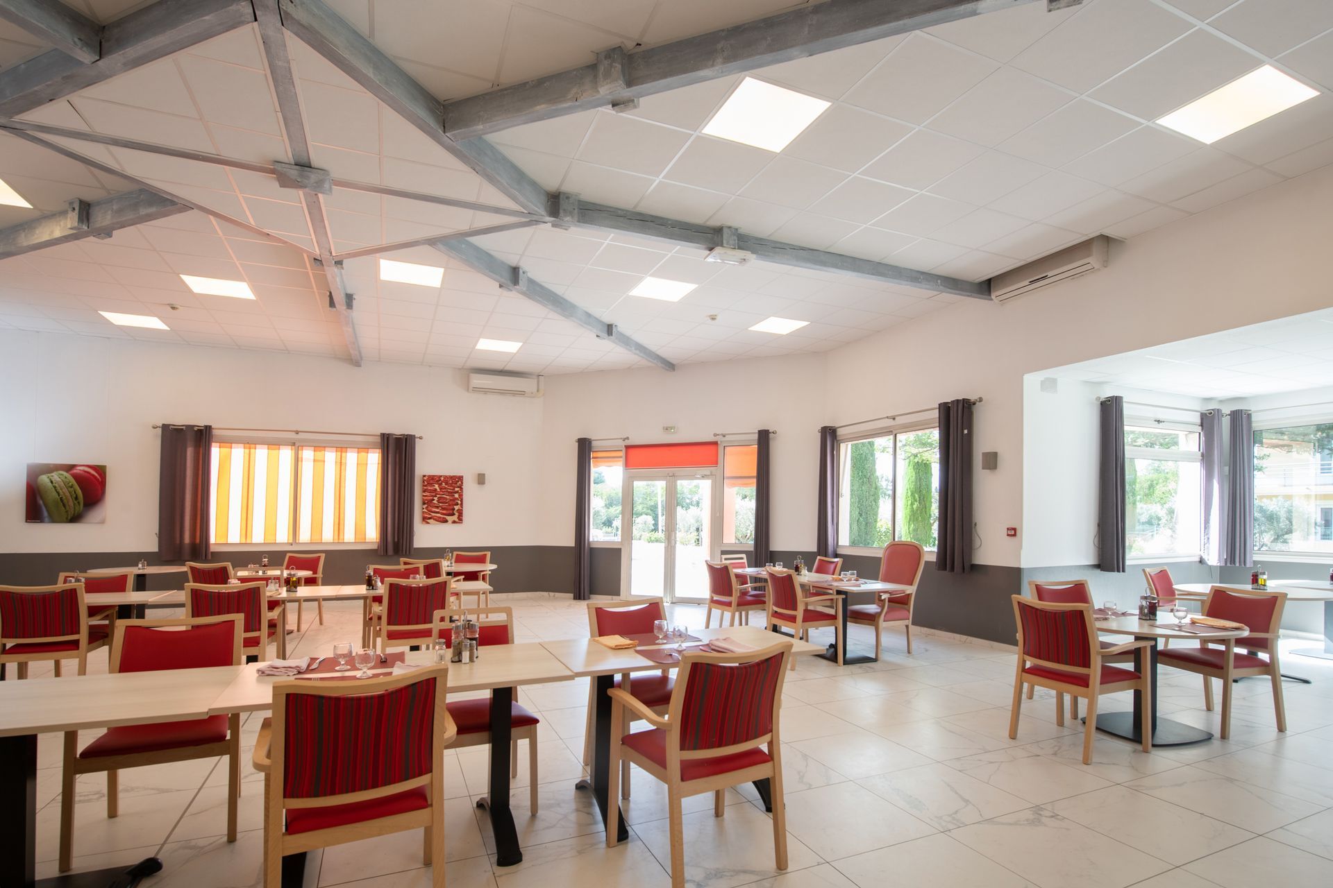Salle à manger lumineuse et moderne avec chaises rouges, tables blanches et grandes fenêtres.