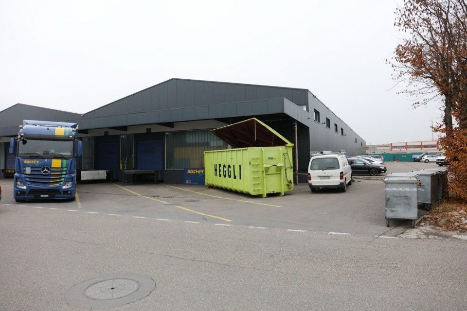 Ein LKW und mehrere Lieferwagen parken an der Laderampe eines Lagerhauses, davor steht ein grüner Müllcontainer.