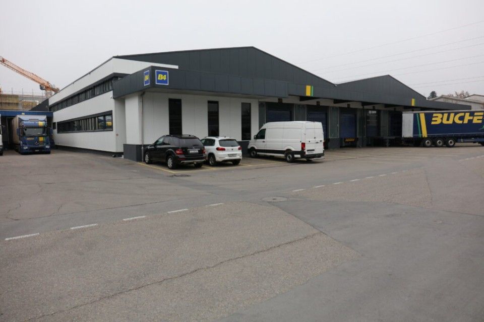 Lagerhalle mit geparkten Autos, einem Lieferwagen und einem Lkw. Aussenansicht, grauer Asphalt, bedeckter Himmel.