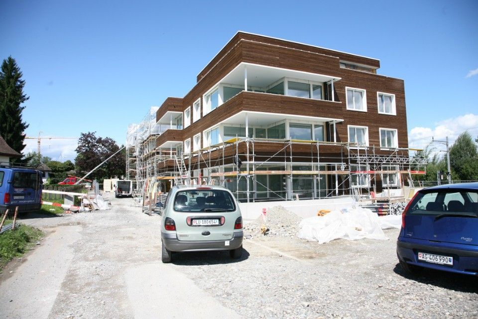 Ein im Bau befindliches Gebäude mit Gerüsten, brauner Fassade, Balkonen, auf der Schotterstrasse geparkten Autos, blauer Himmel.