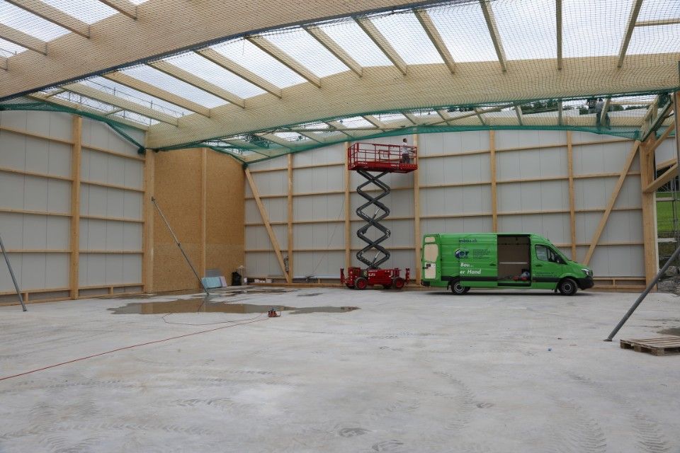 In einem grossen, unfertigen Gebäude mit Holzbalken befinden sich eine Scherenhebebühne und ein grüner Lieferwagen.