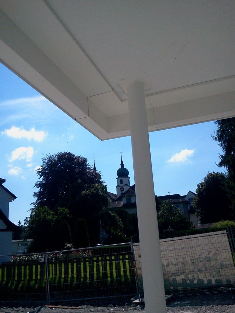 Eine weisse Markise rahmt den Blick auf einen Kirchturm und Bäume vor einem strahlend blauen Himmel ein.
