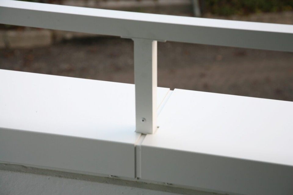 Detail eines weissen Geländers mit vertikaler Stütze und horizontalem oberen Holm an einer niedrigen Mauer.