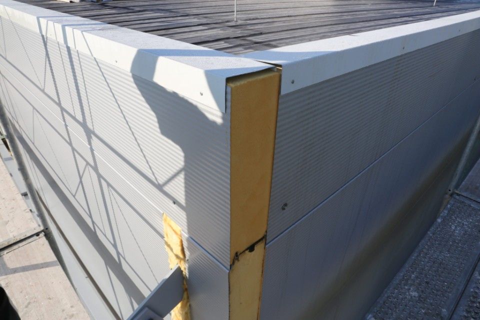 Gebäudeecke mit hellgrauer, gerippter Metallverkleidung und weisen Zierleisten; freiliegende gelbe Isolierung.
