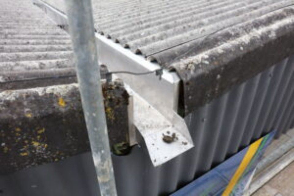 Eine Aluminium-Dachrinne mit Halterung ist an der Kante eines Wellblechdaches befestigt, daneben befinden sich etwas Moos und eine Gerüststange.