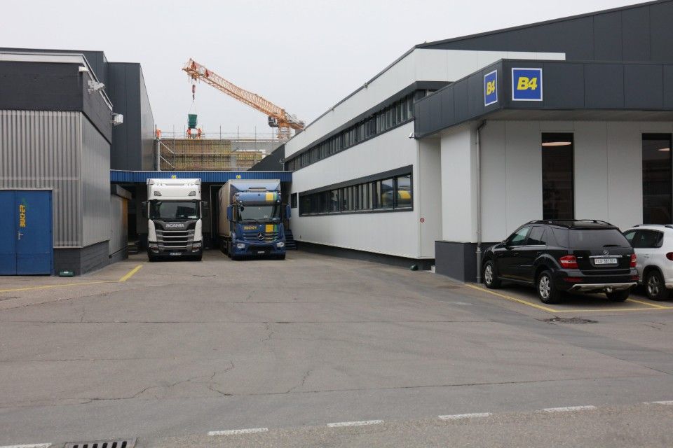 Lastwagen parken vor einem Gebäude mit dem Schild „Bä“, im Hintergrund ein Kran.