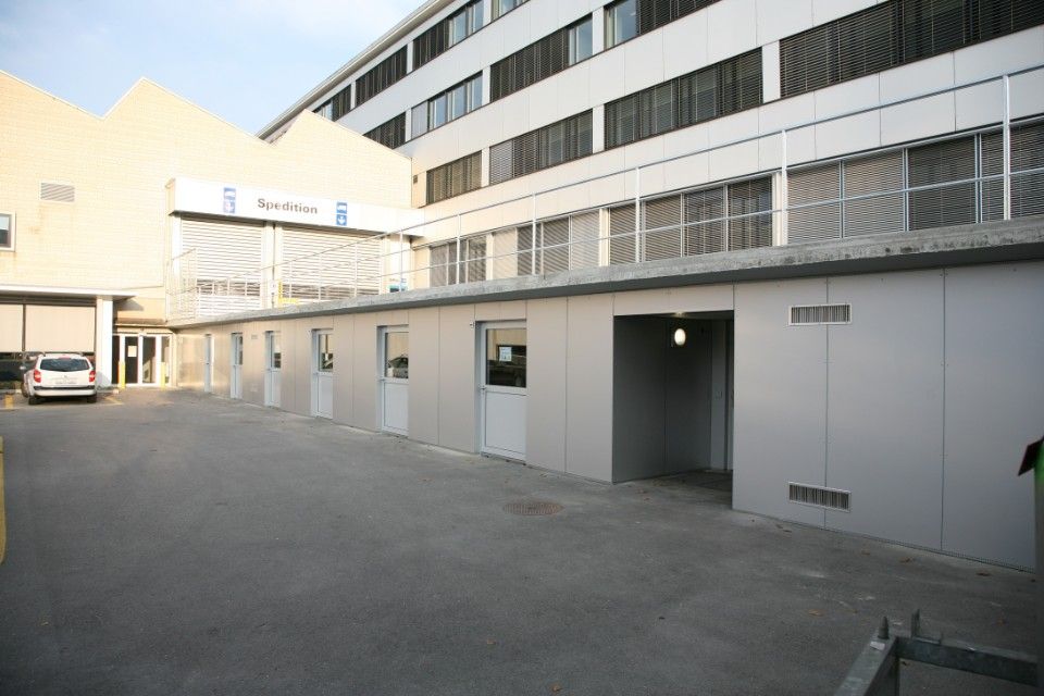 Aussenansicht eines langen, eingeschossigen Modulgebäudes mit einem modernen mehrgeschossigen Gebäude im Hintergrund.