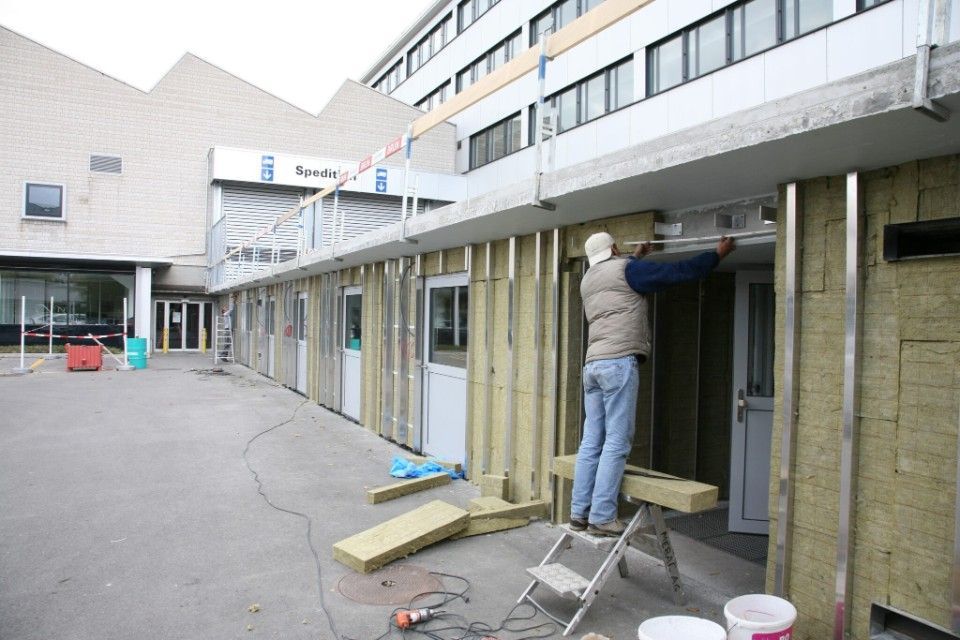 Ein Bauarbeiter bringt Dämmmaterial an einer Aussenwand eines Gebäudes an. Im Hintergrund ist die graue und weisse Gebäudefassade zu sehen.