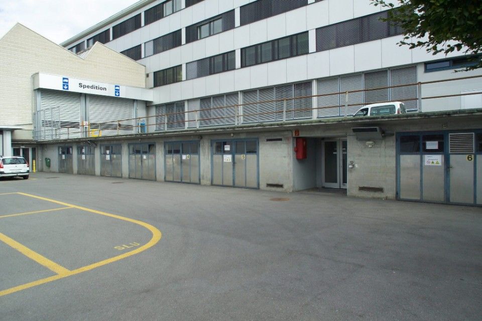 Aussenansicht eines modernen Gebäudes mit Parkplatz, unterhalb der Fenster befinden sich Reihen von Garagentoren. Links ist ein Auto geparkt.