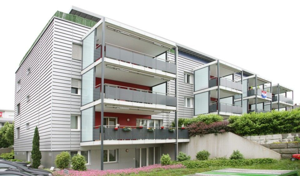 Modernes Mehrfamilienhaus mit Balkonen, grauer Fassade, roten Akzentwänden und Gartenanlage.