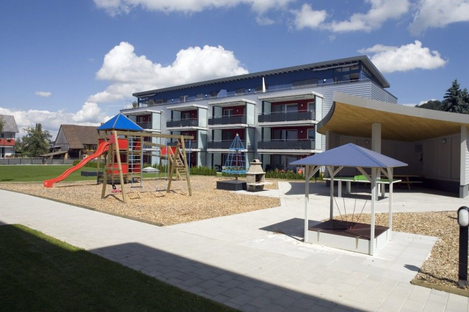 Spielplatz mit Rutschen, Schaukeln und Sandkasten vor einem modernen Wohnhaus unter blauem Himmel.