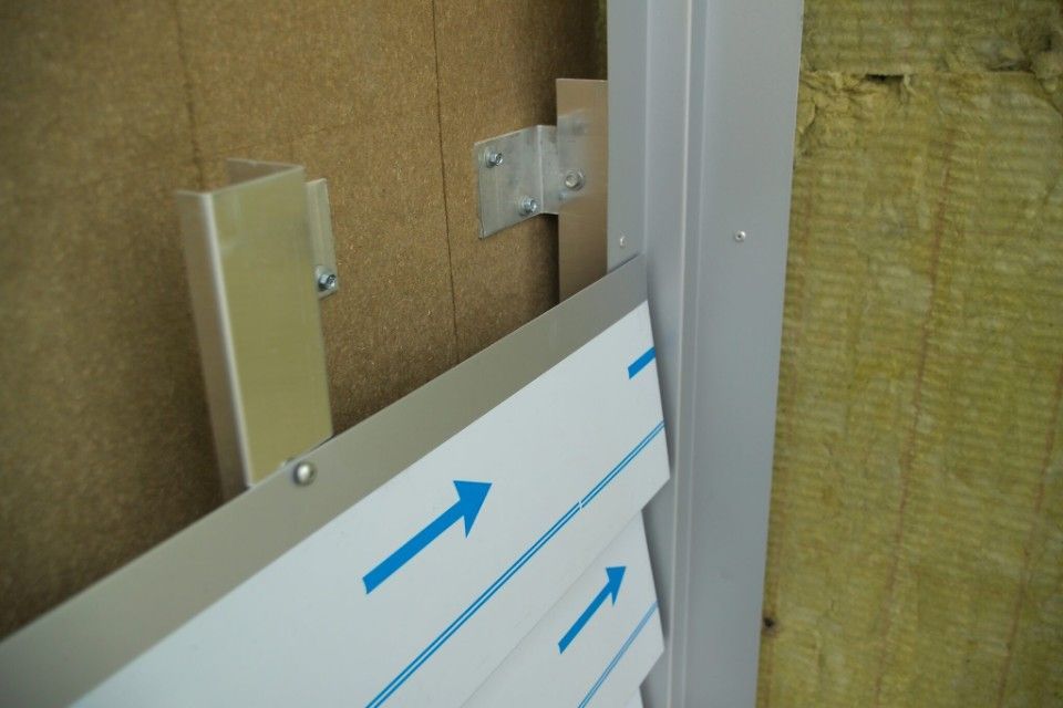 Montage von Aluminium-Fassadenverkleidungen. Die mit blauen Pfeilen gekennzeichneten Verkleidungsteile werden an Metallhalterungen an der Gebäudewand befestigt.