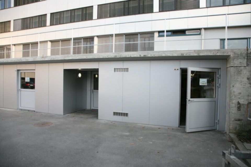 Aussenansicht eines Gebäudes mit mehreren Türen, weisser Metallverkleidung und einer Betonterrasse.