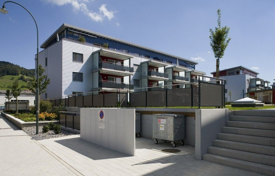 Modernes Mehrfamilienhaus mit Balkonen, Treppen und Müllbehältern im Erdgeschoss.