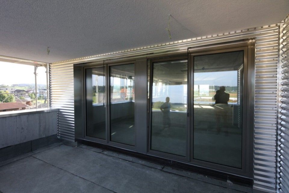 Balkon mit grossen Glasschiebetüren und Wellblechverkleidung. Menschen spiegeln sich im Glas.