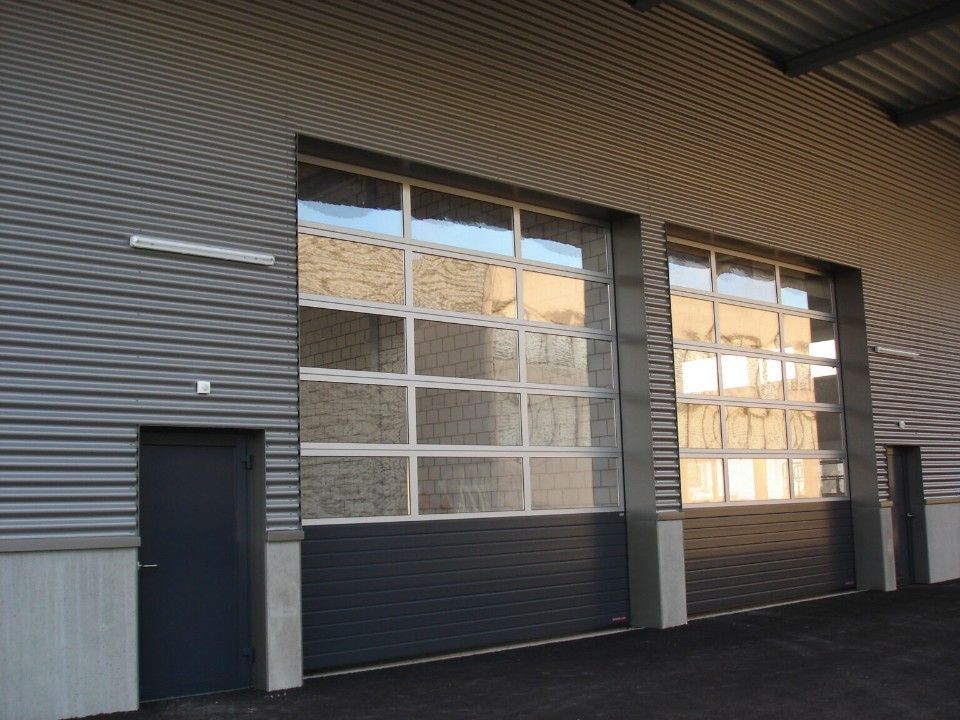 Aussenansicht eines Gebäudes mit zwei grossen Garagentoren mit Fenstern und einer kleinen Tür auf der linken Seite.