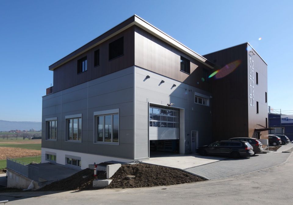 Modernes Industriegebäude mit braunen und grauen Paneelen, Garagentor offen, Autos geparkt, sonniger Tag.