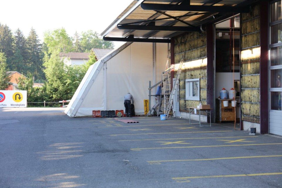 Menschen in der Nähe eines weissen Zeltes und eines Gebäudes mit offenen Garagentoren; asphaltierter Parkplatz.